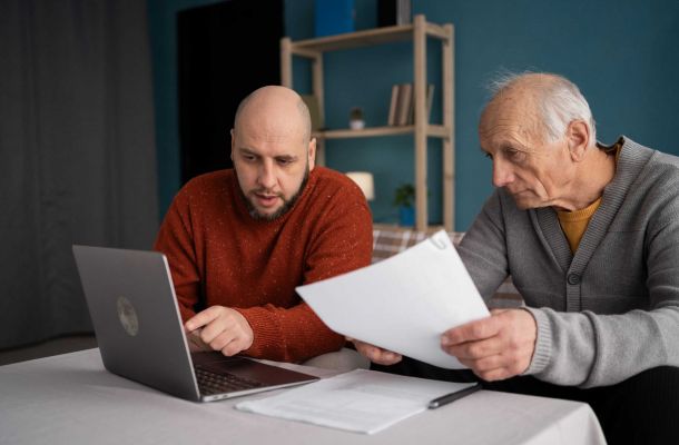 Sohn erklärt seinem älteren Vater am Laptop wichtige finanzielle Unterlagen zum Thema Elternunterhalt.