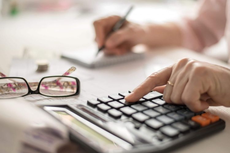 Hand einer Person tippt auf einem Taschenrechner, daneben liegen Unterlagen und eine Brille – symbolisch für die Berechnung von Pflegekosten und finanzielle Planung bei Betreuung.