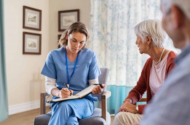 Pflegefachkraft im Gespräch mit älterer Frau bei einem Beratungstermin zu Hause, dokumentiert Informationen in einem Notizbuch