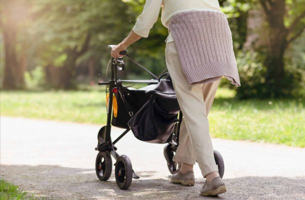 Ältere Frau geht mit einem Rollator auf einem sonnigen Weg im Park – Symbol für Mobilität, Selbstständigkeit und Pflegeleistungen im Alter.