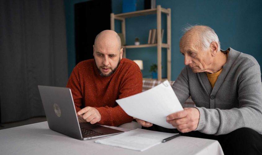 Sohn erklärt seinem älteren Vater am Laptop wichtige finanzielle Unterlagen zum Thema Elternunterhalt.