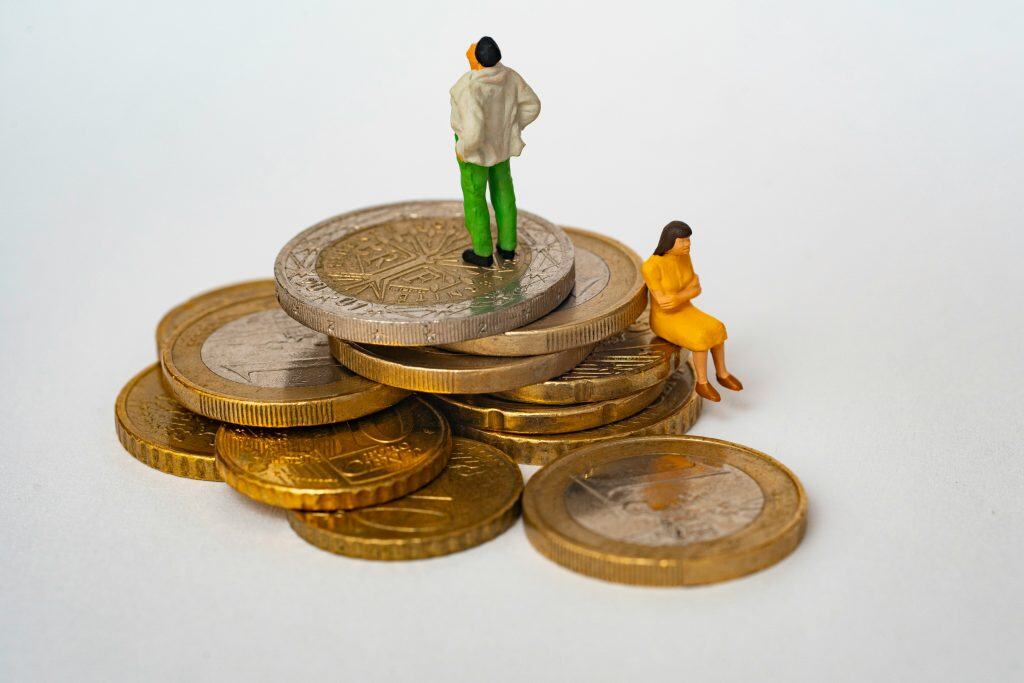 Stapel aus Euro‑Münzen mit zwei kleinen Miniaturfiguren – Symbolbild für steigende Pflegekosten, Mietdruck, häusliche Betreuung und finanzielle Belastung in Ballungsräumen.
