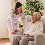 Pflegekraft im Gespräch mit einem älteren Mann in seinem Sessel