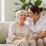 Pflegekraft sitzt mit einer Seniorin auf dem Sofa und hält ihre Hand