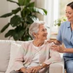 Pflegekraft sitzt neben einer Seniorin auf dem Sofa und legt ihr die Hand auf die Schulter
