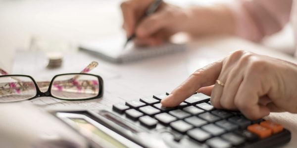 Hand einer Person tippt auf einem Taschenrechner, daneben liegen Unterlagen und eine Brille – symbolisch für die Berechnung von Pflegekosten und finanzielle Planung bei Betreuung.