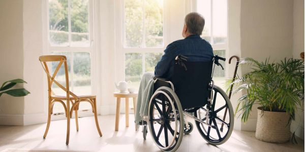 Älterer Mann im Rollstuhl sitzt in seinem Zuhause und blickt nachdenklich aus dem hellen Fenster und will zuhause alt werden.